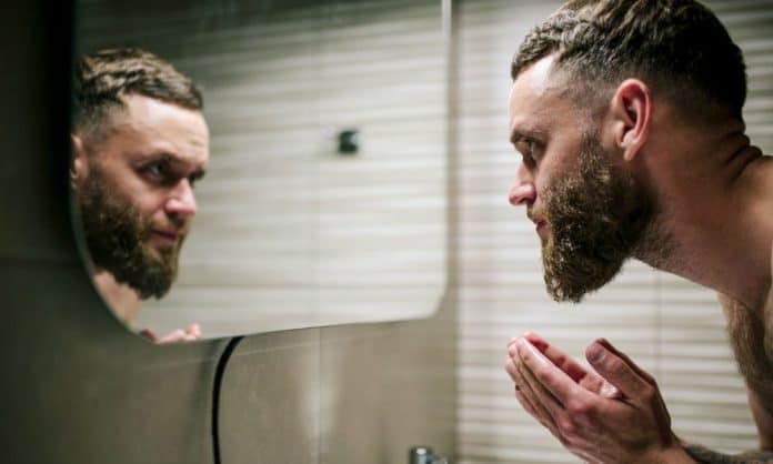 bearded man exfoliates his beard in front of a mirror